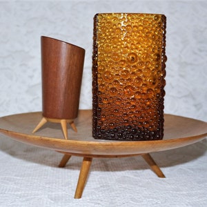 Puede incluir: Una bandeja de madera de estilo moderno de mediados de siglo con tres patas, que sostiene una pequeña taza de madera y un jarrón de vidrio ámbar texturizado. La bandeja y la taza tienen un acabado de madera natural, el jarrón tiene una textura de burbujas.