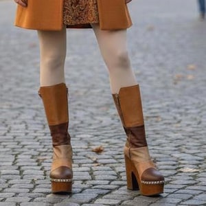 May include: A double-breasted wool coat in a warm amber tone worn over a patterned brown dress with a dark belt. The outfit includes light-colored tights and knee-high platform boots featuring a color-blocked design in shades of brown and tan.