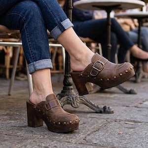 Pode incluir: Tamancos de couro castanho com fivela e detalhes de tachas. Os tamancos têm um salto alto de madeira e sola plataforma. A pessoa está usando jeans azuis com as barras dobradas. Os tamancos são usados em um café ao ar livre.