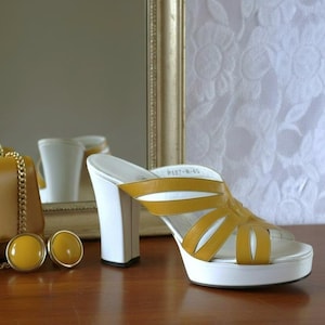 May include: Mustard yellow handbag with a gold chain strap, matching earrings, and a ring. A pair of platform sandals with white soles and heels, and mustard yellow straps are also present. The items are on a wooden surface.