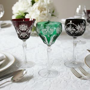 Peut inclure: Verres à vin en cristal de couleur rouge rubis, vert émeraude et noir, avec des motifs floraux et géométriques complexes. Les verres sont posés sur une nappe en dentelle blanche, avec de l'argenterie et des assiettes, suggérant un cadre de restauration formel.