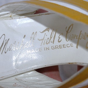 May include: White leather sandals with yellow straps. The sandals have the text "Marshall Field & Company" and "MADE IN GREECE" printed in gold lettering on the insole. The sandals are vintage.