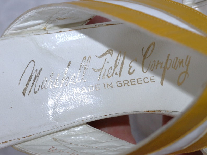 May include: White leather sandals with yellow straps. The sandals have the text "Marshall Field & Company" and "MADE IN GREECE" printed in gold lettering on the insole. The sandals are vintage.