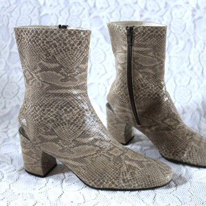 Könnte beinhalten: Ein Paar beige Stiefeletten mit Schlangenmuster. Die Stiefel haben eine quadratische Spitze, einen Blockabsatz und einen Reißverschluss an der Seite. Die Absätze sind ca. 7,6 cm hoch. Auf weißem, strukturiertem Hintergrund.