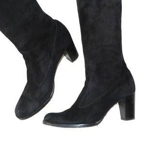 Könnte beinhalten: Ein Paar schwarze Kniehochstiefel aus Wildleder. Die Stiefel haben eine runde Spitze und einen Blockabsatz. Die Absätze sind etwa 7.6 cm hoch. Die Stiefel sind aus einem weichen, samtigen Material gefertigt.