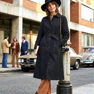 Può includere: Una donna indossa un trench nero e un cappello abbinato, pantaloni di velluto a coste marroni e mocassini con tacco marroni. Una fotocamera vintage è appesa a una cinghia. Scena di strada con auto d'epoca e persone.