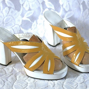 May include: A pair of vintage platform sandals. The sandals feature a mustard yellow and white color scheme. The upper part of the sandal has a unique cut-out design. The heels are thick and measure approximately 4 inches.