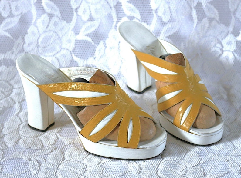 May include: A pair of vintage platform sandals. The sandals feature a mustard yellow and white color scheme. The upper part of the sandal has a unique cut-out design. The heels are thick and measure approximately 4 inches.