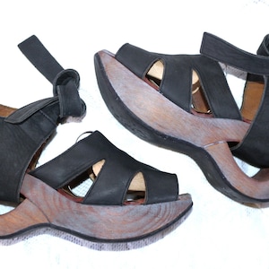 Op de afbeelding: Een paar zwarte leren sandalen met houten plateauzolen. De sandalen hebben een band die om de enkel gaat en een gespsluiting.