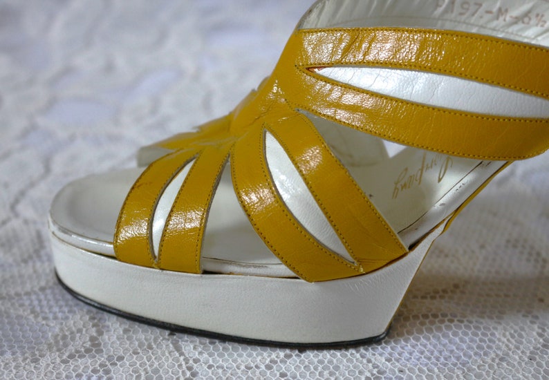 May include: Vintage platform sandals with a white sole and a mustard yellow upper. The open-toe design features cut-out details and a high heel. The sandals have a retro aesthetic, perfect for a vintage-inspired look.