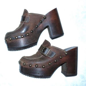 Könnte beinhalten: Ein Paar braune Lederclogs mit Plateau-Sohle und hohem Blockabsatz. Die Clogs haben einen Riemen mit Schnalle und sind mit kleinen, runden Nieten verziert. Die Schuhe haben ein Vintage-Aussehen.