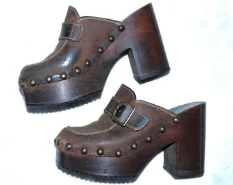 Könnte beinhalten: Ein Paar braune Lederclogs mit Plateau-Sohle und hohem Blockabsatz. Die Clogs haben einen Riemen mit Schnalle und sind mit kleinen, runden Nieten verziert. Die Schuhe haben ein Vintage-Aussehen.