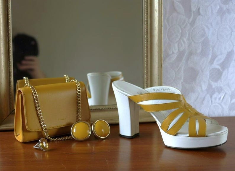 May include: Mustard yellow handbag with a gold chain strap, matching earrings, and a ring. A pair of platform sandals with white soles and heels, and mustard yellow straps are also present. The items are on a wooden surface.