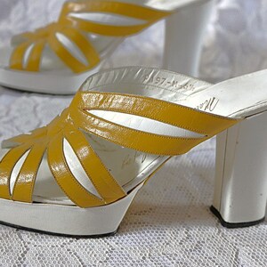 May include: Pair of vintage platform sandals. The open-toe shoes feature a white platform sole and a thick white heel. The upper straps are a bright yellow, with a cut-out design. The sandals have a retro style.