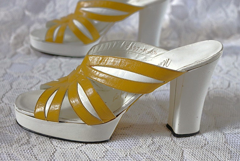 May include: Pair of vintage platform sandals. The open-toe shoes feature a white platform sole and a thick white heel. The upper straps are a bright yellow, with a cut-out design. The sandals have a retro style.
