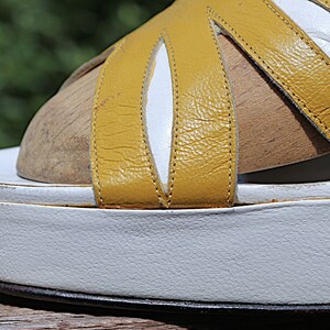 May include: Close-up of a vintage platform sandal. The sandal features a white sole and a yellow leather upper with white accents. The design includes multiple straps and a wooden footbed.