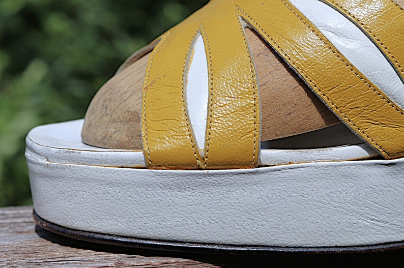 May include: Close-up of a vintage platform sandal. The sandal features a white sole and a yellow leather upper with white accents. The design includes multiple straps and a wooden footbed.