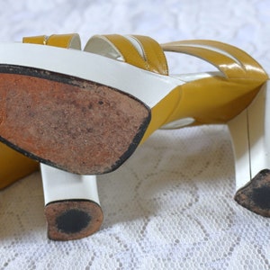 May include: A pair of vintage platform sandals. The sandals feature a white base with mustard yellow straps and a block heel. The soles are worn, showing signs of use. The heels are approximately 3 inches (7.6 cm) high.