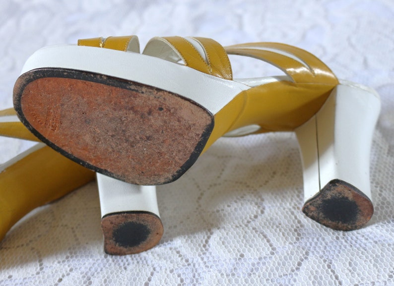 May include: A pair of vintage platform sandals. The sandals feature a white base with mustard yellow straps and a block heel. The soles are worn, showing signs of use. The heels are approximately 3 inches (7.6 cm) high.