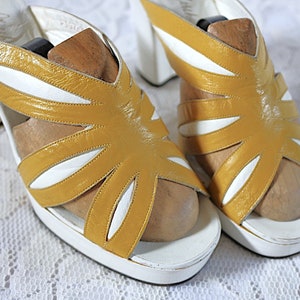 May include: A pair of vintage platform sandals. The sandals feature a mustard yellow and white color scheme with a unique cut-out design. The heels are white and the sandals have wooden shoe forms inside.