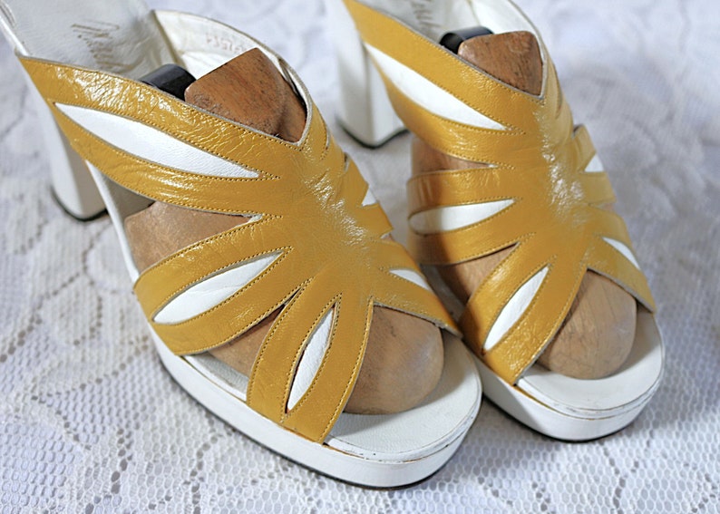 May include: A pair of vintage platform sandals. The sandals feature a mustard yellow and white color scheme with a unique cut-out design. The heels are white and the sandals have wooden shoe forms inside.