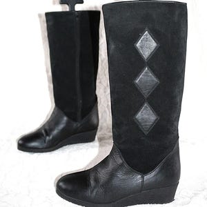 Op de afbeelding: Twee zwarte kniehoge laarzen van leer en suède. De laarzen hebben een sleehak en een decoratief ruitpatroon op de suède schacht. Ze hangen aan zwarte kleerhangers.