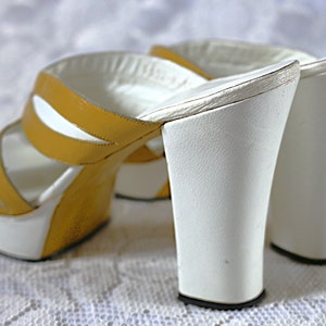 May include: A pair of vintage platform sandals. The open-toe sandals feature a white platform sole and a white, flared heel. The upper straps are mustard yellow with white accents. The sandals are set against a white, textured background.