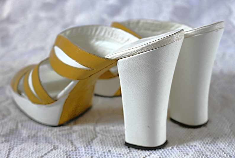 May include: A pair of vintage platform sandals. The open-toe sandals feature a white platform sole and a white, flared heel. The upper straps are mustard yellow with white accents. The sandals are set against a white, textured background.