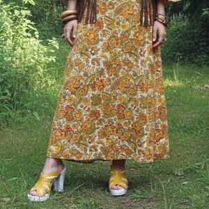 May include: A long, flowing dress with a paisley pattern in shades of yellow, orange, and green. The dress is paired with yellow sandals with white soles and heels. The overall style suggests a bohemian or vintage aesthetic.