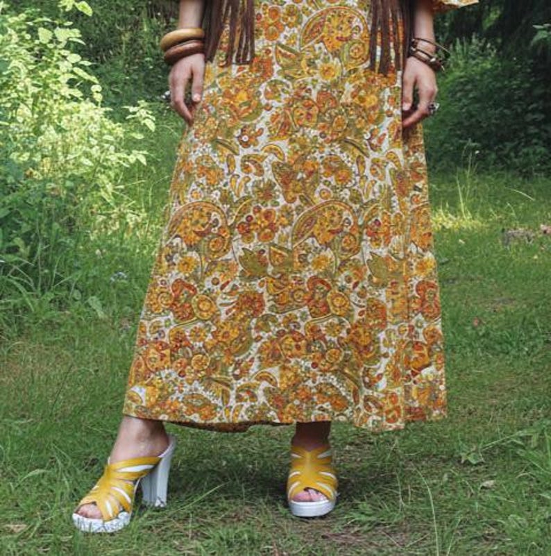 May include: A long, flowing dress with a paisley pattern in shades of yellow, orange, and green. The dress is paired with yellow sandals with white soles and heels. The overall style suggests a bohemian or vintage aesthetic.
