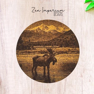 May include: A round wooden coaster with a brown and gold design featuring a moose standing in a lake with mountains in the background. The text "Zen Imperium Designs" is printed at the top of the coaster.