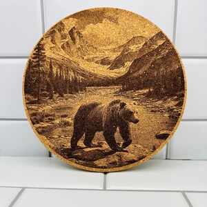 May include: A round cork trivet with a brown bear walking through a mountain landscape. The bear is in the foreground and the mountains are in the background. The trivet is a light brown color.