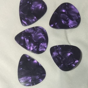 May include: Five purple guitar picks with a pearlescent finish. The picks are shaped like a triangle with rounded edges.