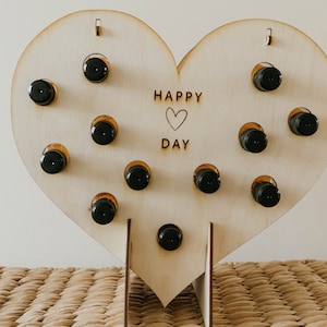 Puede incluir: Soporte de madera en forma de corazón con 12 pequeños porta botellas. El soporte está grabado con el texto "HAPPY ♥ DAY".