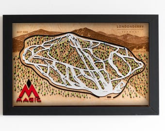 May include: A framed wooden wall art piece depicting a 3D topographic ski trail map of Magic Mountain in Londonderry, Vermont. The map features white raised ski trails, green trees, and the red Magic logo on a light wood background in a black frame.