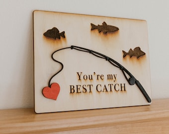 Puede incluir: Señal de madera con un corazón rojo, una caña de pescar y tres peces. El letrero dice "You're my BEST CATCH".