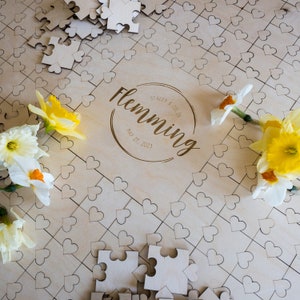 May include: A wooden heart-shaped puzzle guest book with the names Stacey & Colin and the text "Flemming" and "May 27, 2023" engraved on it. The puzzle is surrounded by yellow daffodils.