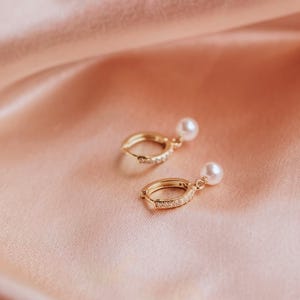 May include: A pair of gold-colored hoop earrings with a small white pearl dangling from each. The hoops are embellished with small, clear stones. The earrings rest on a pink satin surface. Text at the bottom reads "6mm pearls".