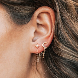 May include: Gold-colored earrings featuring celestial designs. One earring is a starburst with a dangling chain, and the other is a crescent moon. The earrings are worn on an ear, with the person's hair visible in the background.
