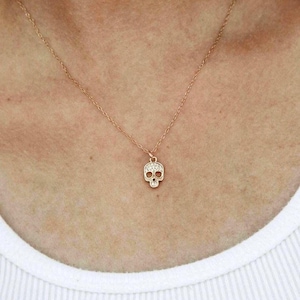 May include: A delicate gold-colored necklace featuring a small skull pendant. The skull is adorned with small, sparkling crystals. The necklace is displayed against a white background.
