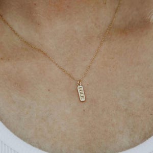 May include: A gold-colored necklace with a delicate chain. The pendant is a small, rectangular bar with the word "FUCK" engraved in a vertical orientation. The necklace is worn against a person's skin.