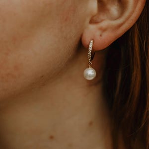 May include: Gold hoop earrings with a single pearl dangling from each hoop. The hoops are set with small clear stones.