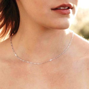 May include: A delicate silver chain necklace with a simple, elongated oval link design. The necklace is worn around the neck, creating a minimalist and elegant look. The chain is thin and appears to be made of a shiny metal.