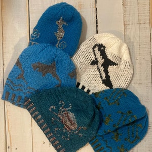 May include: A collection of knitted beanies in various shades of blue, teal, and white. Each hat features a unique design, including seahorses, sharks, anchors, and a stylized penguin. The hats are arranged on a white wooden surface.
