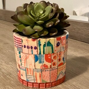 Op de afbeelding: Een kleine succulent plant in een kleurrijke mozaïek pot. De pot is gemaakt van kleine tegels in tinten roze, oranje, geel, blauw en groen.