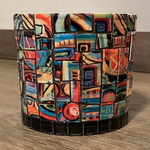 Pode incluir: Um vaso de mosaico colorido com um padrão geométrico. O vaso é feito de pequenos azulejos quadrados em tons de azul, vermelho, amarelo, verde e preto. O vaso tem uma borda preta.