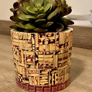 Pode incluir: Um pequeno vaso cilíndrico feito de mosaicos com uma variedade de cores e padrões. O vaso está cheio de uma planta suculenta verde.