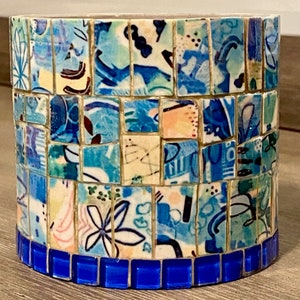 Op de afbeelding: Een cilindrische keramische container met een mozaïekontwerp. De mozaïek is gemaakt van kleine, kleurrijke tegels in tinten blauw, wit en geel. De container heeft een blauwe rand rond de bovenrand.
