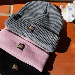 Puede incluir: Tres gorros de punto en gris, rosa y negro, apilados. Cada gorro tiene una pequeña etiqueta negra con un logotipo de corona dorada. Un grupo de cristales de cuarzo transparente está al lado.