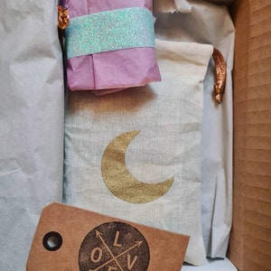 Könnte beinhalten: Eine kleine, weiße Kordeltasche mit einem goldenen Halbmond-Design. Die Tasche befindet sich in einem Karton mit einem in rosa Seidenpapier verpackten Geschenk und einem braunen Etikett mit dem Wort "LOVE" in einem Kreis mit gekreuzten Pfeilen.